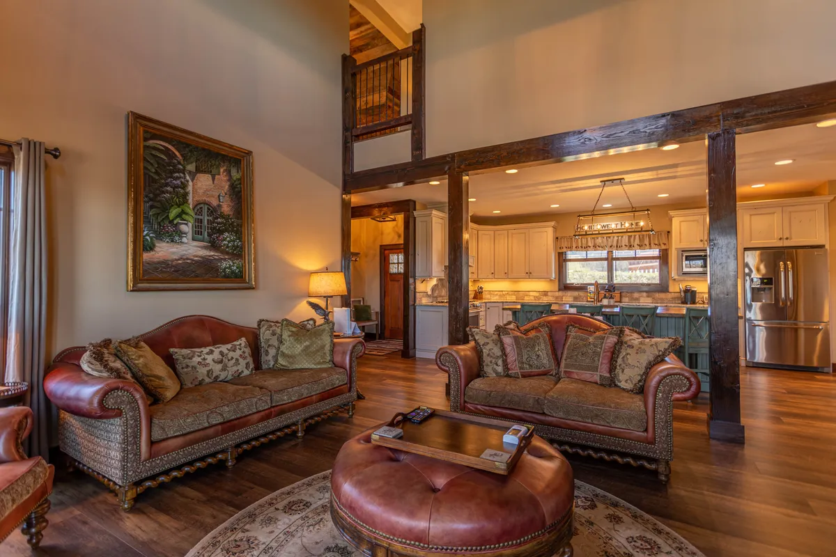 An open, comfortable layout with soft sofas, exposed wooden beams, a visible kitchen area in the background, and tasteful decor. The space flows naturally from the living area into the dining and kitchen zones, encouraging conversation and shared experiences.