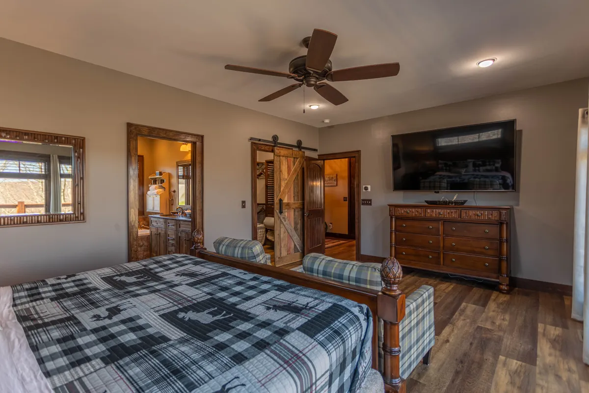 This image showcases a bedroom with a large bed dressed in plaid bedding, a spacious wooden dresser, a wall-mounted television, and a rustic barn-style sliding door. Warm, natural light enhances the cozy and inviting atmosphere, offering a glimpse of the room’s relaxing ambiance and entertainment amenities.