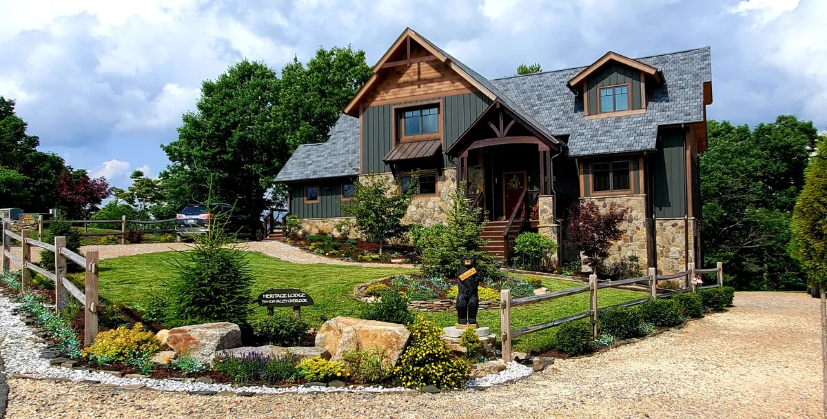 The image shows the facade of a beautiful, newly built cabin nestled on a lush mountainside in Banner Elk, North Carolina. A charming stone pathway leads through a landscaped garden to the welcoming porch, perfect for enjoying sunset views or a quiet morning coffee. The natural surroundings and expansive outdoor space set a tranquil, inviting mood.