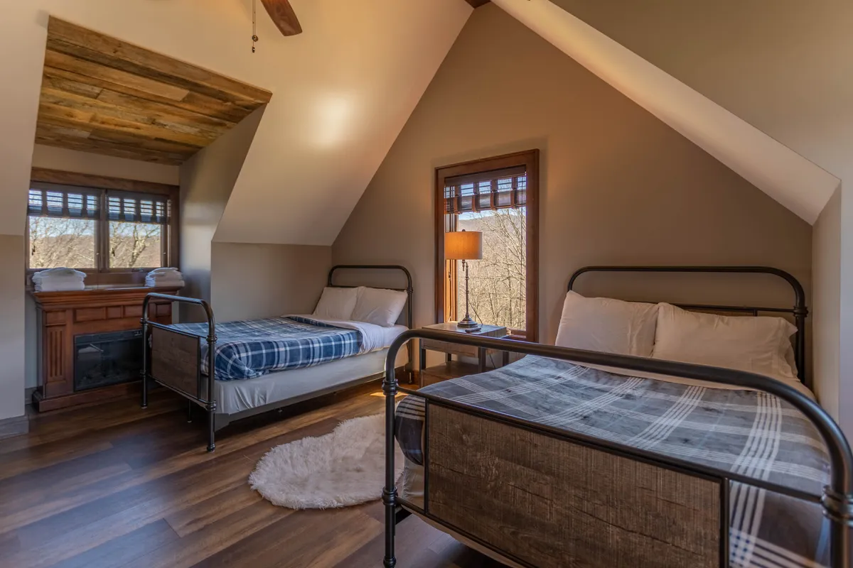 This inviting bedroom features two beds with plaid bedding, a wooden ceiling, large windows that let in natural light, and a small fireplace, creating a warm and serene space for rest or reading. The setup is ideal for families or friends, offering both comfort and charm.
