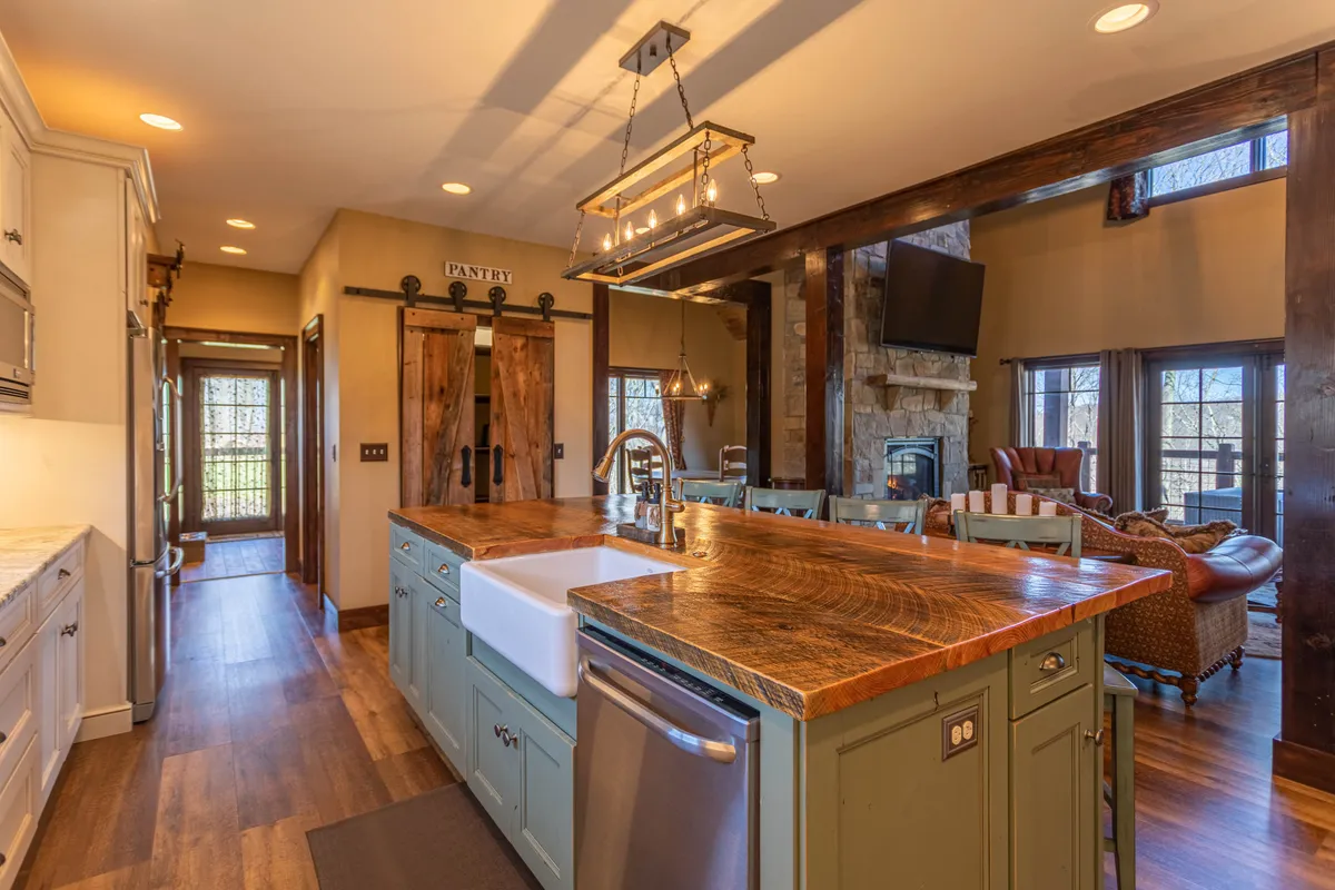 A cozy kitchen area featuring a prominent island, wooden beams, pantry doors, large windows, and a nearby fireplace. The space is flooded with natural light and designed for gathering, cooking, and relaxed evenings.