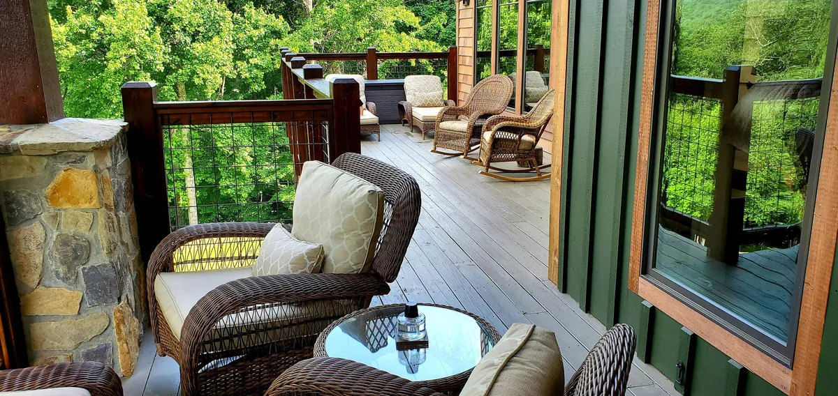 A cozy exterior seating area with wicker chairs and a glass table, set against a backdrop of wooden railing and lush greenery. The setup is perfect for socializing, reading, or simply soaking in the peaceful natural surroundings.