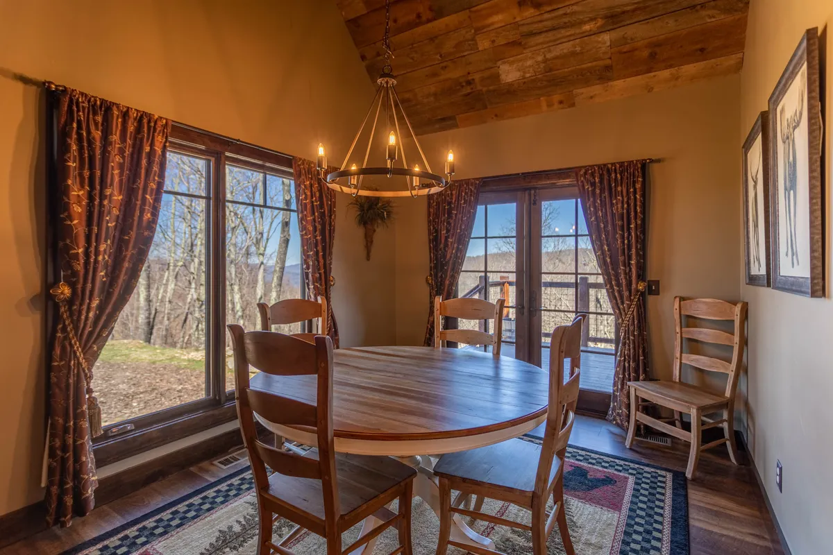 A warm and welcoming dining area featuring a round wooden table, a prominent chandelier, and large windows with curtains. This space is set for family meals, intimate gatherings, or wine tastings, offering plenty of natural light and a convivial atmosphere.