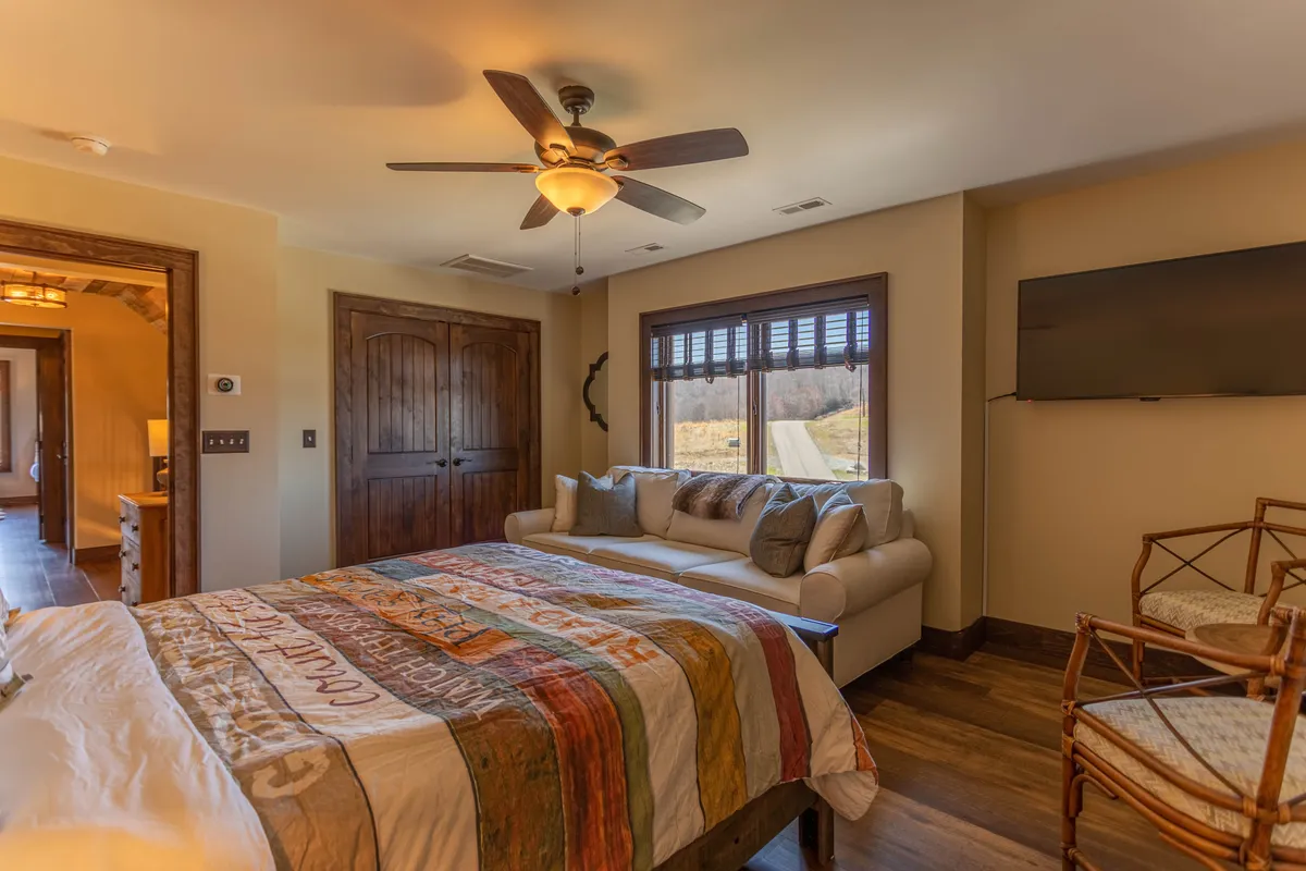 A bedroom scene featuring a bed with a colorful quilt, a comfy sofa with cushions, wooden doors, and a window with scenic views. The space exudes warmth and flexibility, offering both a sleeping area and a place to relax or watch TV, suitable for different guest needs.